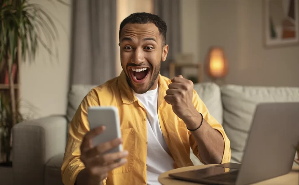 Anunciar Imóveis na Internet: A imagem mostra um homem sentado em um ambiente moderno, provavelmente uma sala de estar ou escritório. Ele está sorrindo e parece estar em um momento de alegria ou satisfação. O homem está usando um telefone celular e parece estar olhando para a câmera, possivelmente tirando uma selfie. Ele está vestindo uma jaqueta amarrada e uma camiseta branca por baixo. Ao lado dele, há um laptop aberto, sugerindo que ele pode estar trabalhando ou desfrutando de algum tipo de entretenimento digital. O ambiente é iluminado e aconchegante, com uma parede decorada e uma planta ao fundo.