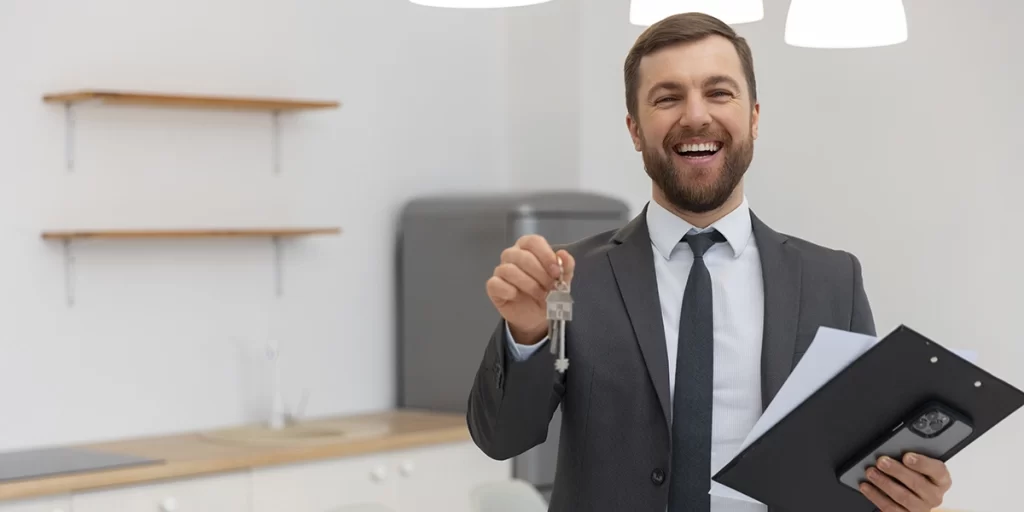 Apresentação do Corretor de Imóveis: A imagem mostra um homem em um ambiente que parece ser uma oficina ou sala de trabalho. Ele está vestindo um terno preto e uma gravata preta, e está sorrindo para a câmera. O homem está segurando uma chave de um edifício e um livro ou folha de papel. Ele parece estar feliz e confiante, possivelmente em um momento de sucesso ou realização profissional.