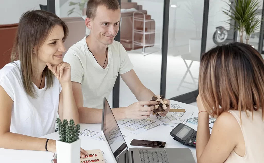 Como Captar Clientes para a Venda de Imóveis: A imagem mostra três pessoas sentadas em volta de uma mesa, parecendo estar em um ambiente de trabalho ou de negociações. Um homem está de pé, sorrindo e segurando algo que parece ser um modelo ou um objeto de design. Ele está vestindo uma camiseta branca e parece estar falando ou apresentando algo. Duas mulheres estão sentadas, uma delas está olhando para ele com uma expressão de interesse, enquanto a outra está olhando para a mesa, possivelmente lendo ou analisando algo. A mesa está comum, com um laptop aberto, um celular, um cálculador e algumas outras coisas que parecem ser documentos ou planos. Há uma planta decorativa no canto superior direito da imagem. O ambiente é moderno e iluminado, com uma decoração minimalista.