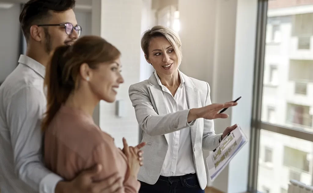 Como Captar Clientes para a Venda de Imóveis: A imagem mostra três pessoas em um ambiente interno, provavelmente uma oficina ou sala de negociações. Uma mulher está segurando um caderno e parece estar apresentando ou discutindo algo com os outros dois indivíduos, que estão de pé e olhando para ela. A mulher está vestida com um terno claro e parece estar em um papel de liderança ou apresentação. Os outros dois estão vestidos com camisas brancas e parecem estar atentos à apresentação. A sala tem uma janela grande com vista para outros edifícios, sugerindo que está em um local urbano.