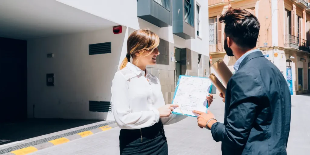 Captação de imóveis Captação de imóveis: imagem mostra uma mulher segurando uma planilha e mostrando a um homem. Ao fundo da imagem há uma casa de dois andares e um imóvel com arquitetura antiga.