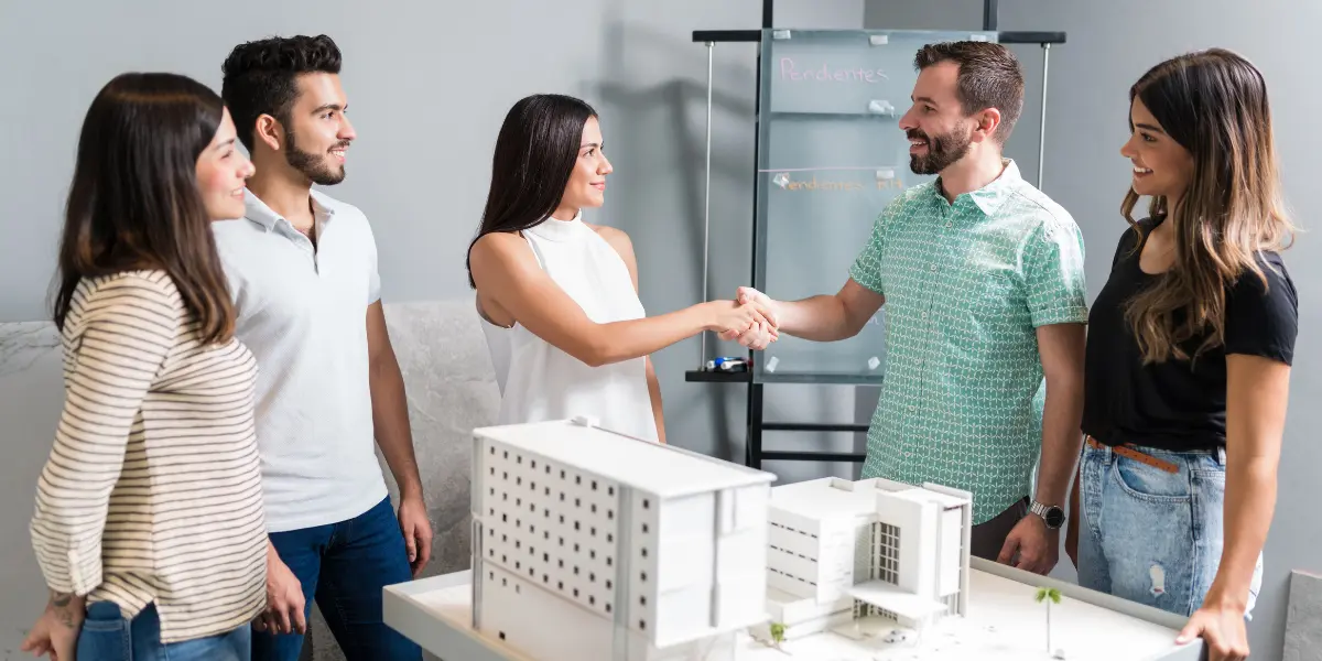 Lançamentos imobiliários: Vários indivíduos estão reunidos em uma sala, possivelmente um escritório de arquitetura, observando um modelo de edifício em uma mesa.
