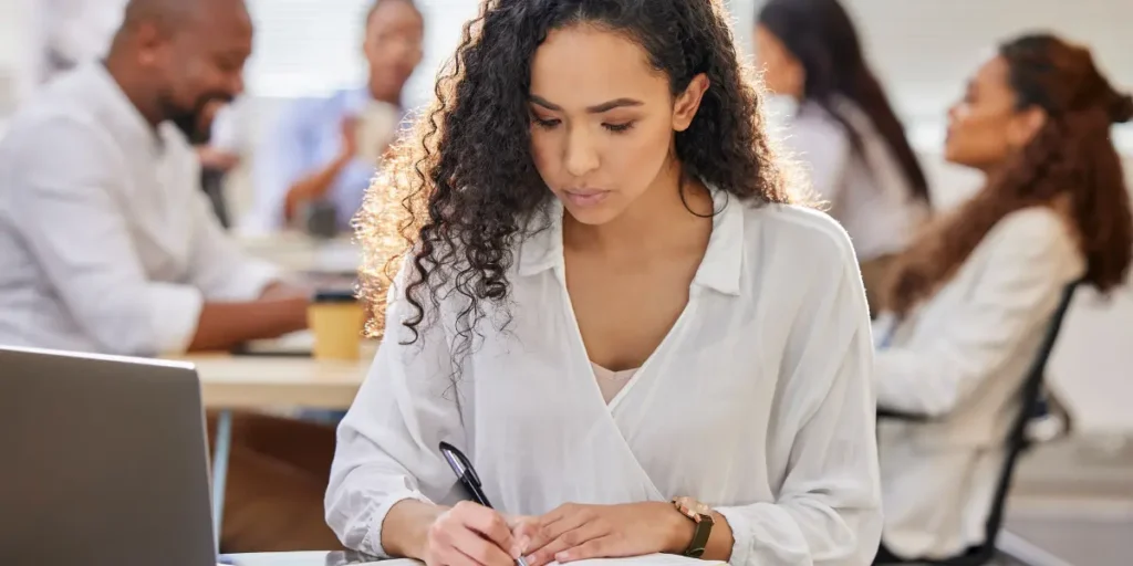Curso TTI: A composição da imagem foca em primeiro plano em uma mulher jovem sentada à mesa, segurando uma caneta anotando, e com um notebook a frente. Em segundo plano há outras pessoas desfocadas, sentadas em uma mesa redonda. 