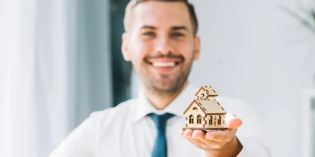 Curso TTI: um homem em segundo plano, desfocado, segura uma miniatura de casa em madeira, em primeiro plano.