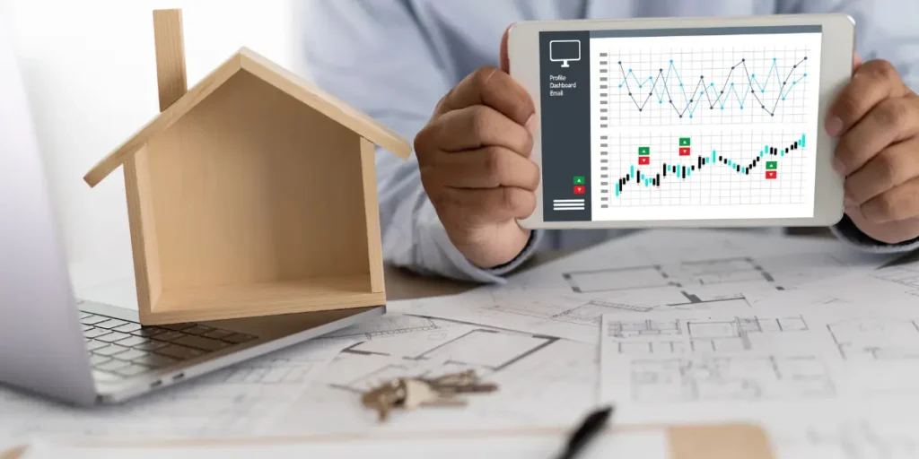 Precificação de imóveis Precificação de imóveis: A composição da cena apresenta a parte superior de uma pessoa segurando um tablet, exibindo um painel de controle com gráficos, posicionando-se ao centro. No lado esquerdo, há uma pequena casa de madeira colocada sobre um laptop. Ao fundo, espalhada sobre a mesa, encontram-se plantas e chaves, sugerindo um ambiente de trabalho relacionado a imóveis.