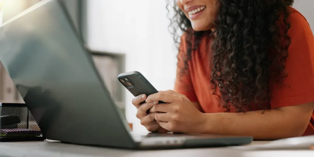 pagadoria online: pagadoria online: mulher segura um celular com as duas mãos e sorri olhando para a tela. Na frente dela há um notebook.
