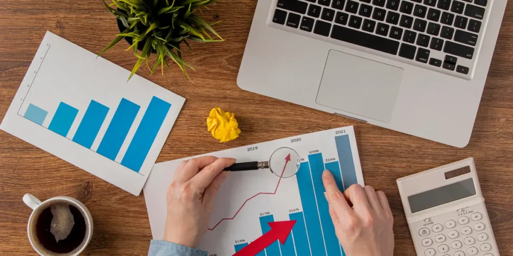 processo comercial em incorporadoras: A composição da cena é organizada de forma simétrica, com o laptop no canto superior direito, uma planta no topo central, e gráficos e outros acessórios distribuídos harmoniosamente. A cena mostra a ação de alguém analisando os gráficos, com as mãos segurando uma lupa e apontando para os dados.