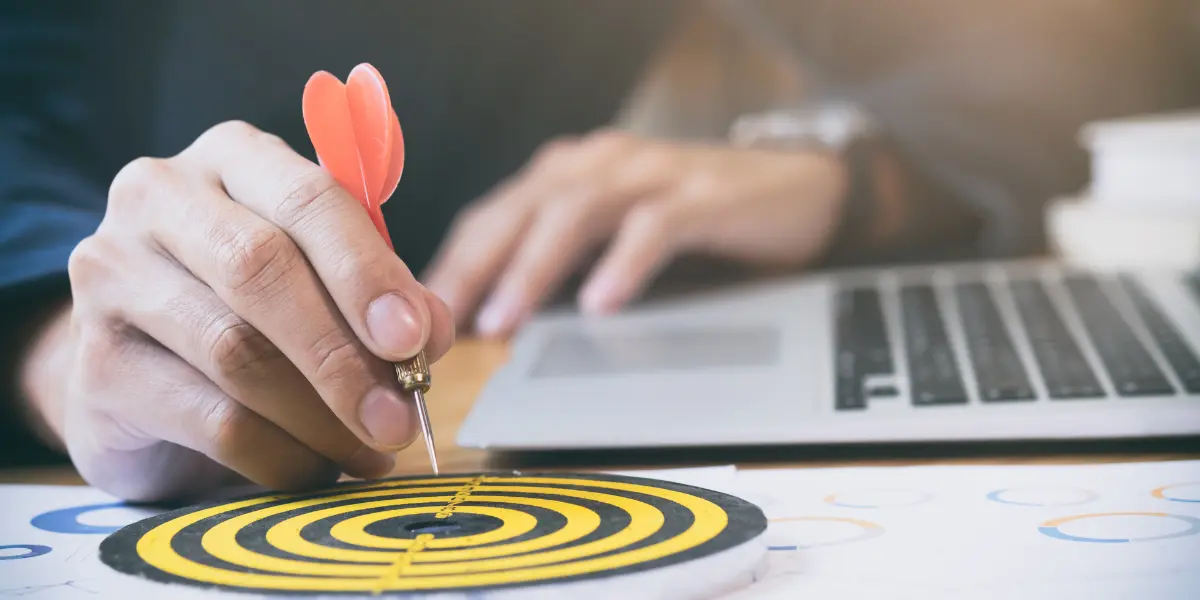 processo comercial em incorporadoras: a imagem mostra uma mão segurando o dardo que aponta para o centro do alvo, situado na parte inferior da imagem. O restante da cena é preenchido com um laptop aberto ao fundo e papéis com gráficos espalhados na superfície, sugerindo um ambiente de trabalho.
