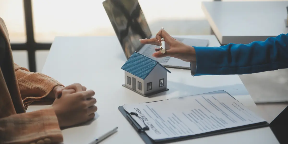 CPF do imóvel: A composição mostra uma mesa branca, com uma modelo de casa no centro e papéis de contrato na frente. Uma pessoa, possivelmente um corretor, gesticula com a mão direita, apontando para o modelo da casa, enquanto outra pessoa sentada em frente observa. Um laptop aberto e canetas complementam o cenário,