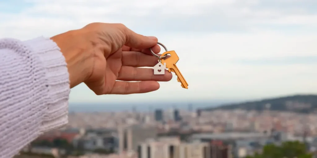 CPF do imóvel: A fotografia mostra uma mão segurando um molho de chaves de metal dourado com um chaveiro em forma de casa, tendo uma cidade desfocada como fundo.