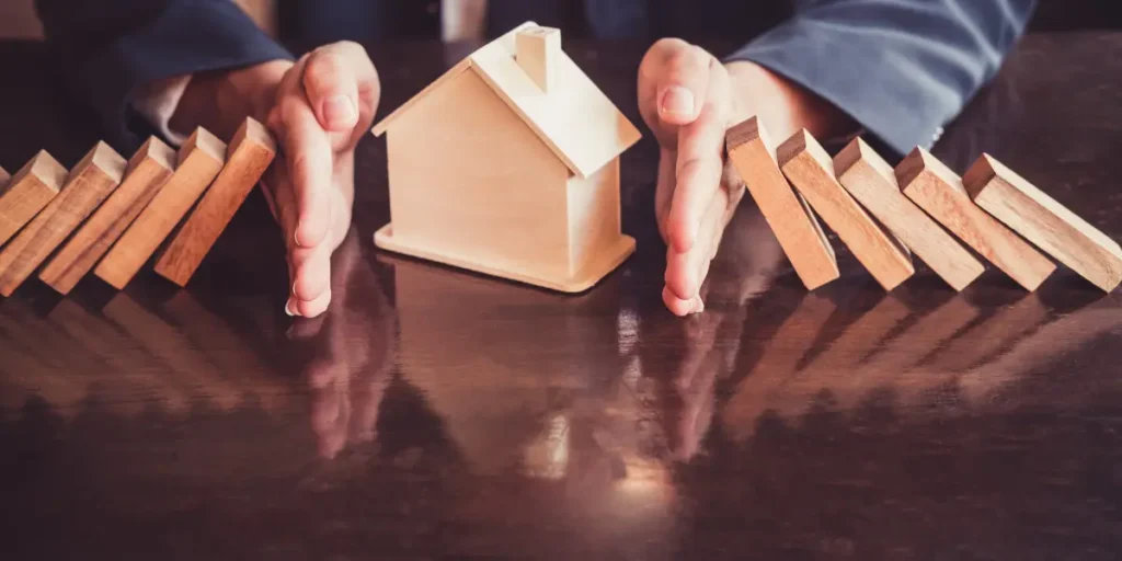 comissão imobiliária comissão imobiliária: a imagem mostra mãos centralizadas e uma casa em miniatura posicionada entre elas. Há blocos de madeira, dispostos em fila, que estão prestes a cair.