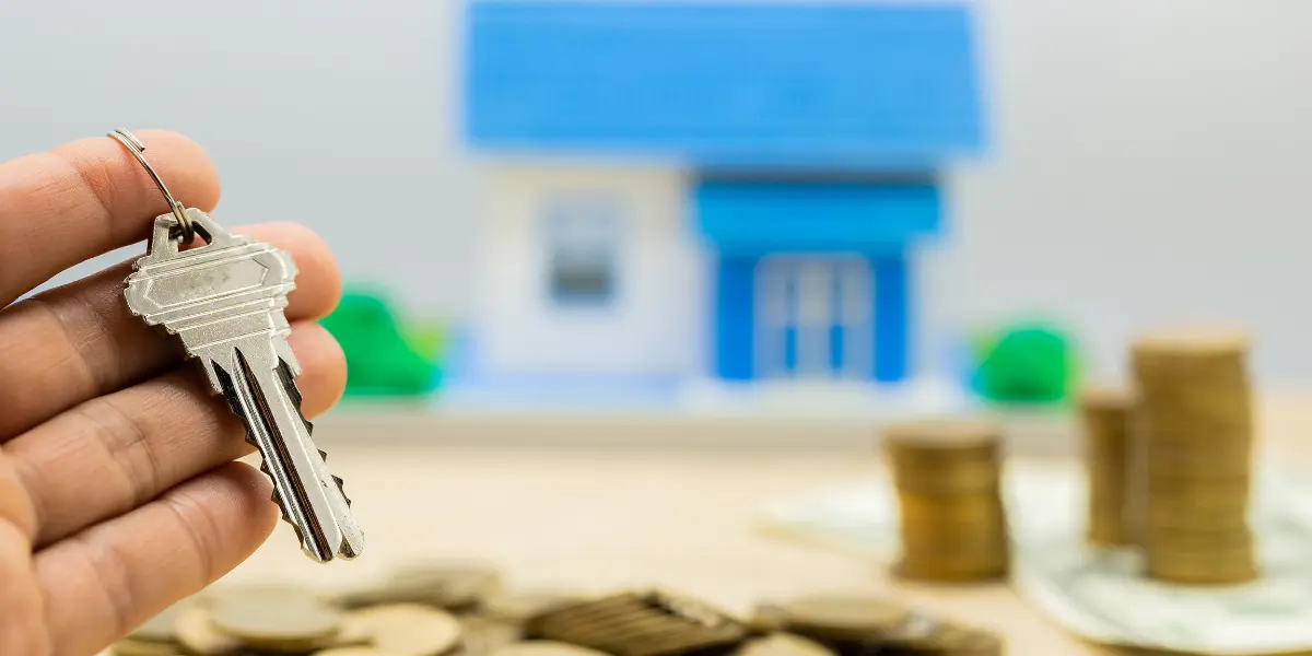 comissão imobiliária: foto apresenta uma mão segurando uma chave de casa com uma casa em miniatura borrada e pilhas de moedas ao fundo.