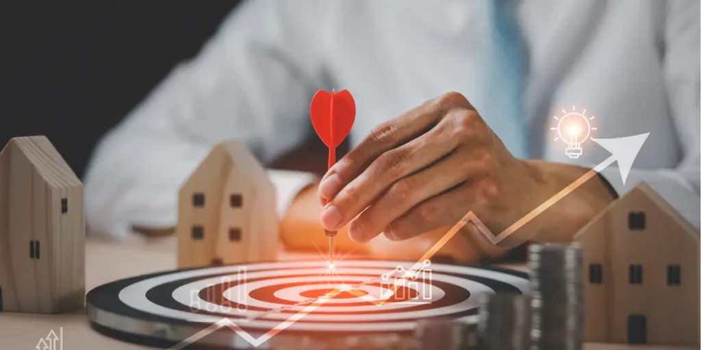 Pós-venda no mercado imobiliário Pós-venda no mercado imobiliário: A cena é focada na mão de uma pessoa, segurando uma seta vermelha com precisão, apontando para o centro de um alvo de dardos. Ao redor do alvo, há casas de madeira em miniatura, moedas empilhadas e gráficos de linha ascendentes e um ícone de lâmpada, todos sugerindo conceitos de investimento, planejamento financeiro e crescimento.
