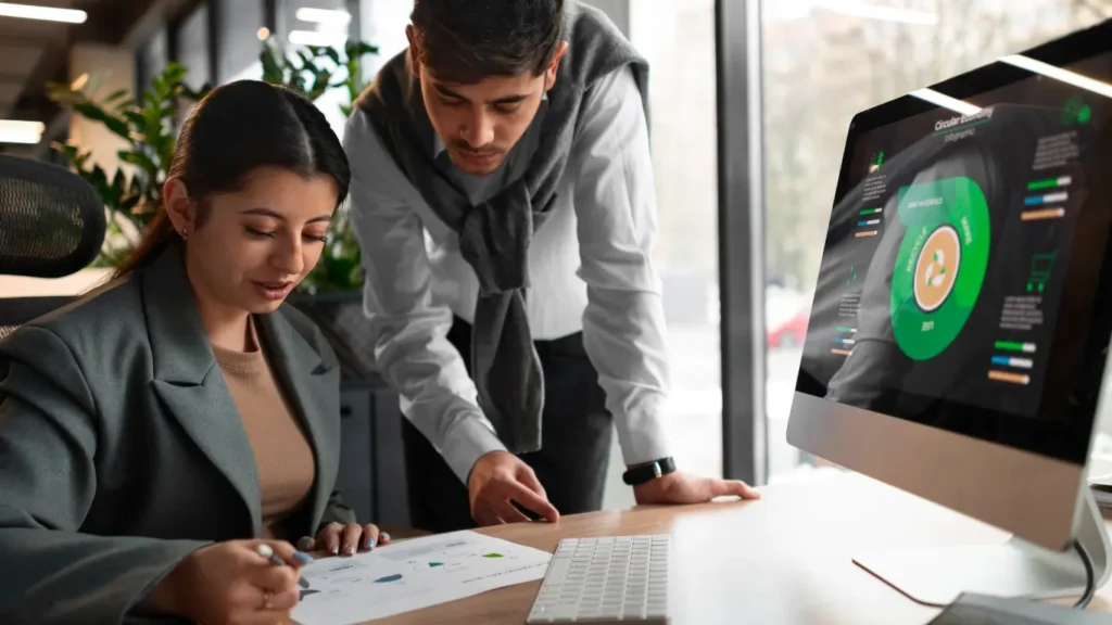 parcerias imobiliárias parcerias imobiliárias: A composição da cena é organizada em torno de uma mesa de escritório. Uma mulher está sentada, inclinada sobre um documento, enquanto um homem está em pé ao lado dela, inclinado sobre a mesa, aparentemente olhando para o mesmo documento ou para o monitor do computador. O computador com tela em um ângulo que mostra um gráfico circular é posicionado à direita. A iluminação é suave, vinda de uma janela à direita, e a ação sugere uma discussão colaborativa.
