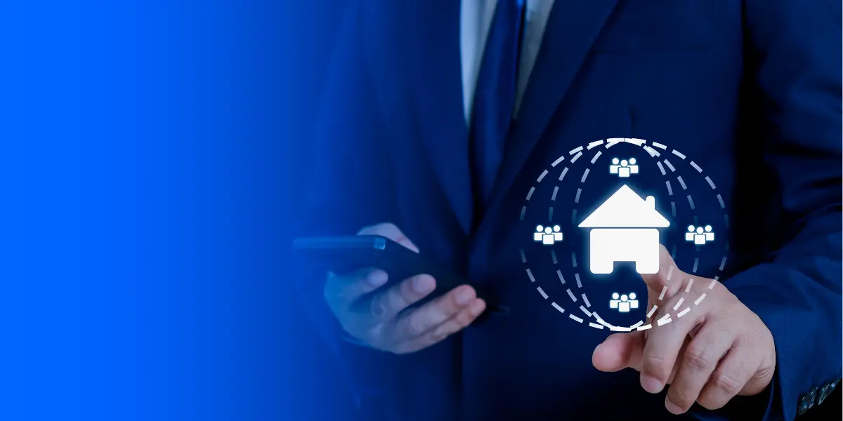 IA para corretores de imóveis: A composição da cena apresenta um close-up parcial de um homem vestido com um elegante terno azul escuro, com as mãos visíveis. Ele segura um smartphone com a mão esquerda e com a mão direita, ele toca um ícone holográfico em forma de casa, cercado por figuras de pessoas e linhas pontilhadas, possivelmente representando uma rede ou conexão. O plano de fundo é predominantemente azul escuro, com um gradiente que sugere profundidade e foco na interação.