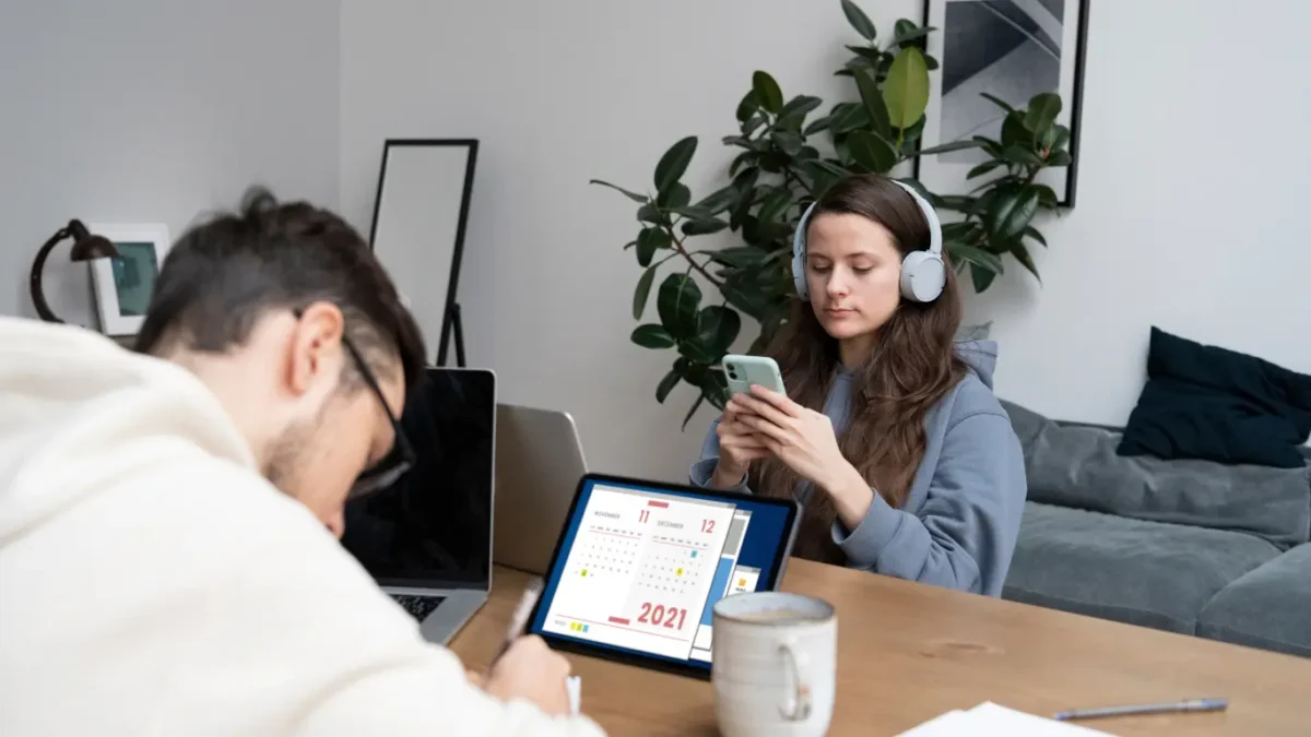 Duas pessoas trabalhando em uma mesa de madeira em ambiente interno; um homem escreve ao lado de um notebook, enquanto uma mulher usa fones de ouvido e mexe no celular, com um tablet exibindo um calendário sobre a mesa e plantas ao fundo. automação de marketing imobiliário