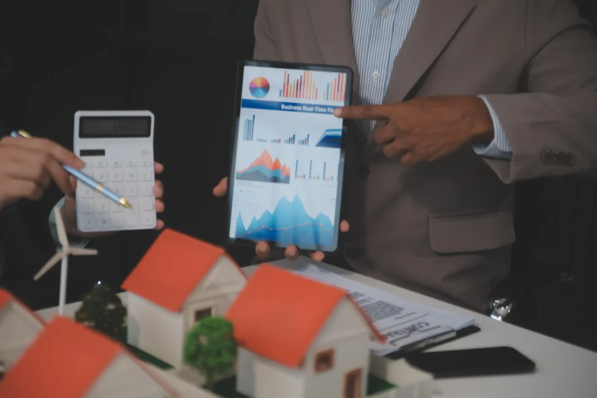 otimização de vendas imobiliárias: A composição da cena demonstra uma reunião de negócios, com duas pessoas interagindo em um ambiente profissional. Um homem, à direita, segura um tablet exibindo gráficos e parece apontar para eles, enquanto outra pessoa, à esquerda, segura uma caneta e uma calculadora, sugerindo uma análise ou negociação financeira.