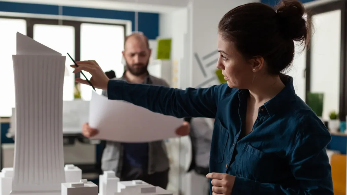 Profissional analisa a maquete de um edifício enquanto aponta detalhes com um lápis, em um ambiente de trabalho com outra pessoa ao fundo segurando plantas do projeto.