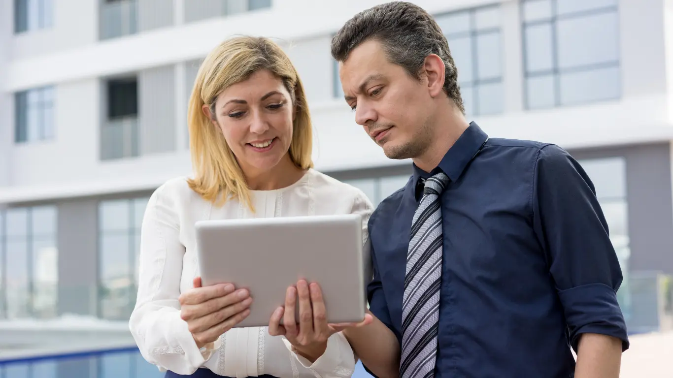 Homem e mulher em ambiente externo, em frente a um prédio moderno, analisando informações em um tablet enquanto conversam.