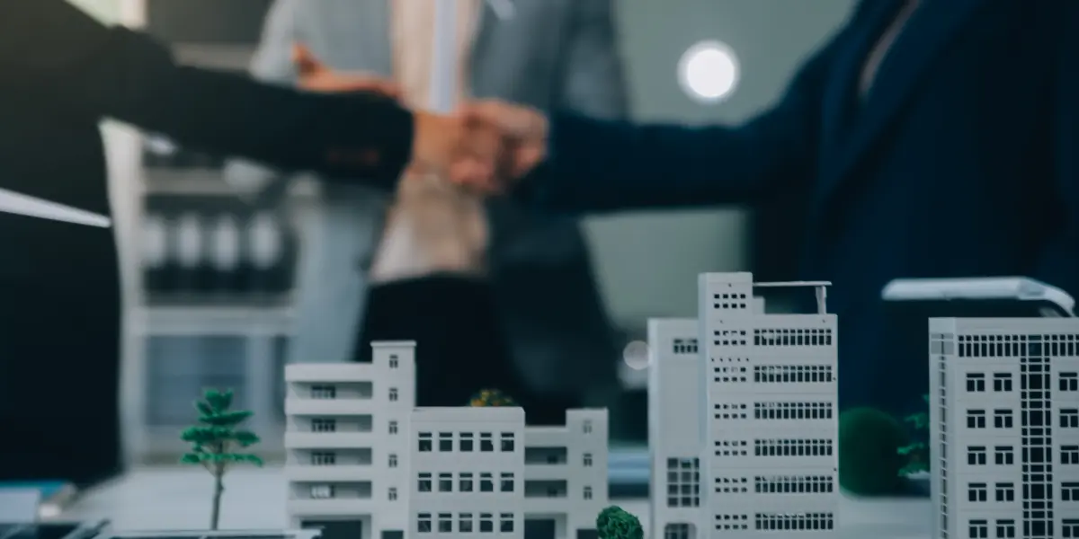 Parcerias imobiliárias estratégicas: A imagem exibe um aperto de mão entre colegas, com modelos de edifícios em primeiro plano, sugerindo um acordo imobiliário.