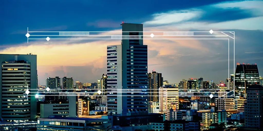 Inovação imobiliária: A composição da cena exibe uma paisagem urbana vasta e densa, dominada pela silhueta de vários edifícios altos. No centro da imagem, um arranha-céu proeminente se destaca, com sua fachada lisa e janelas escuras contrastando com o céu. A imagem está disposta de forma a capturar a extensão da cidade, com os edifícios se estendendo por toda a largura, sugerindo uma metrópole expansiva. Um contorno gráfico em branco, com elementos decorativos geométricos, é sobreposto à imagem.