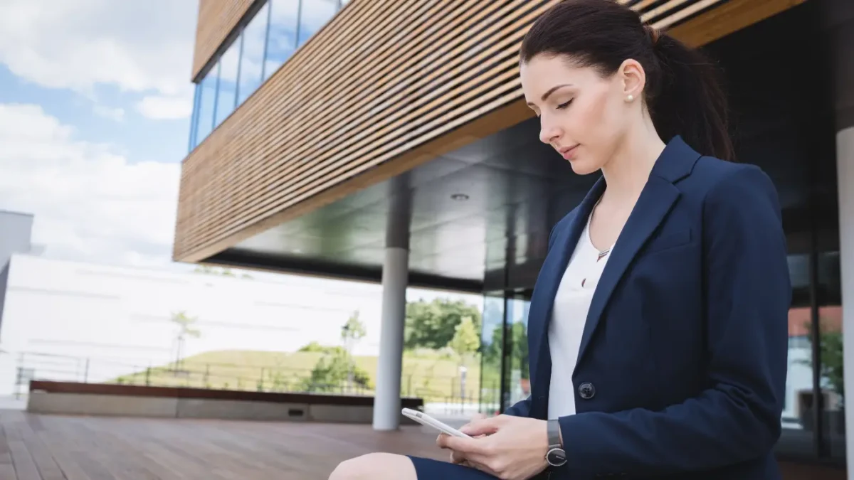 Mulher vestindo blazer azul utilizando smartphone em frente a prédio corporativo moderno com fachada de vidro e madeira.