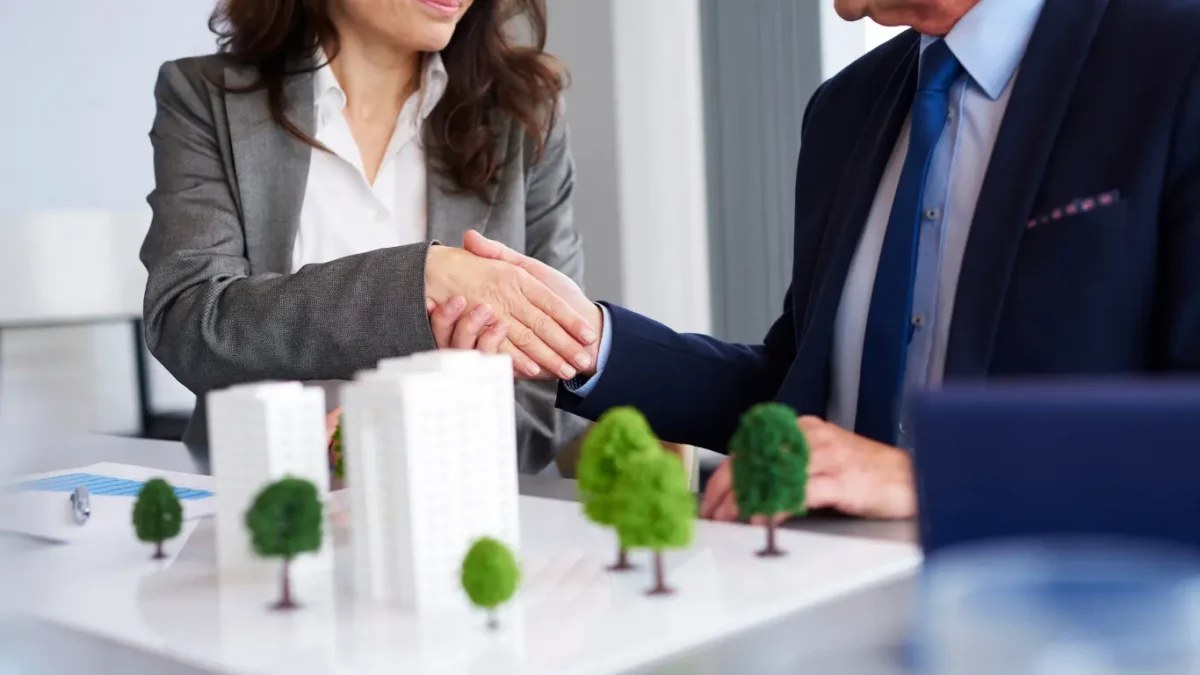 Duas pessoas em traje formal apertando as mãos sobre uma mesa com maquete de empreendimento imobiliário, simbolizando acordo ou fechamento de negócio.