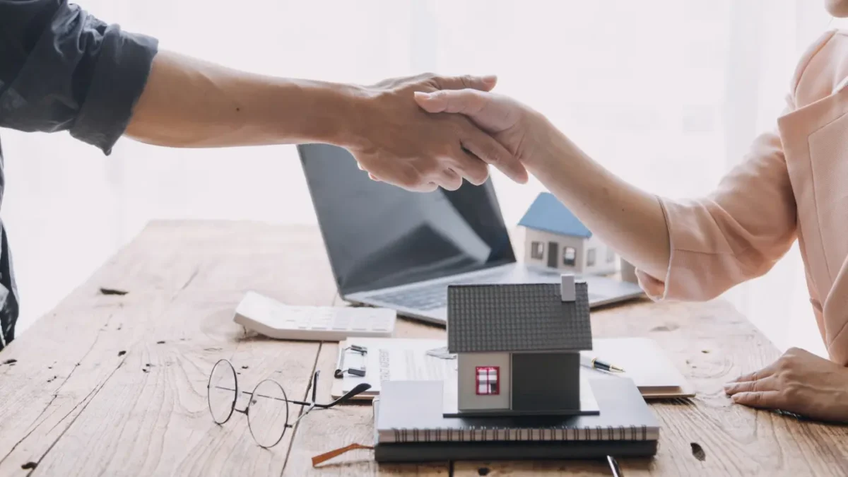 Duas pessoas apertando as mãos sobre uma mesa com uma maquete de casa, documentos e um notebook ao fundo, representando acordo ou negociação no mercado imobiliário.