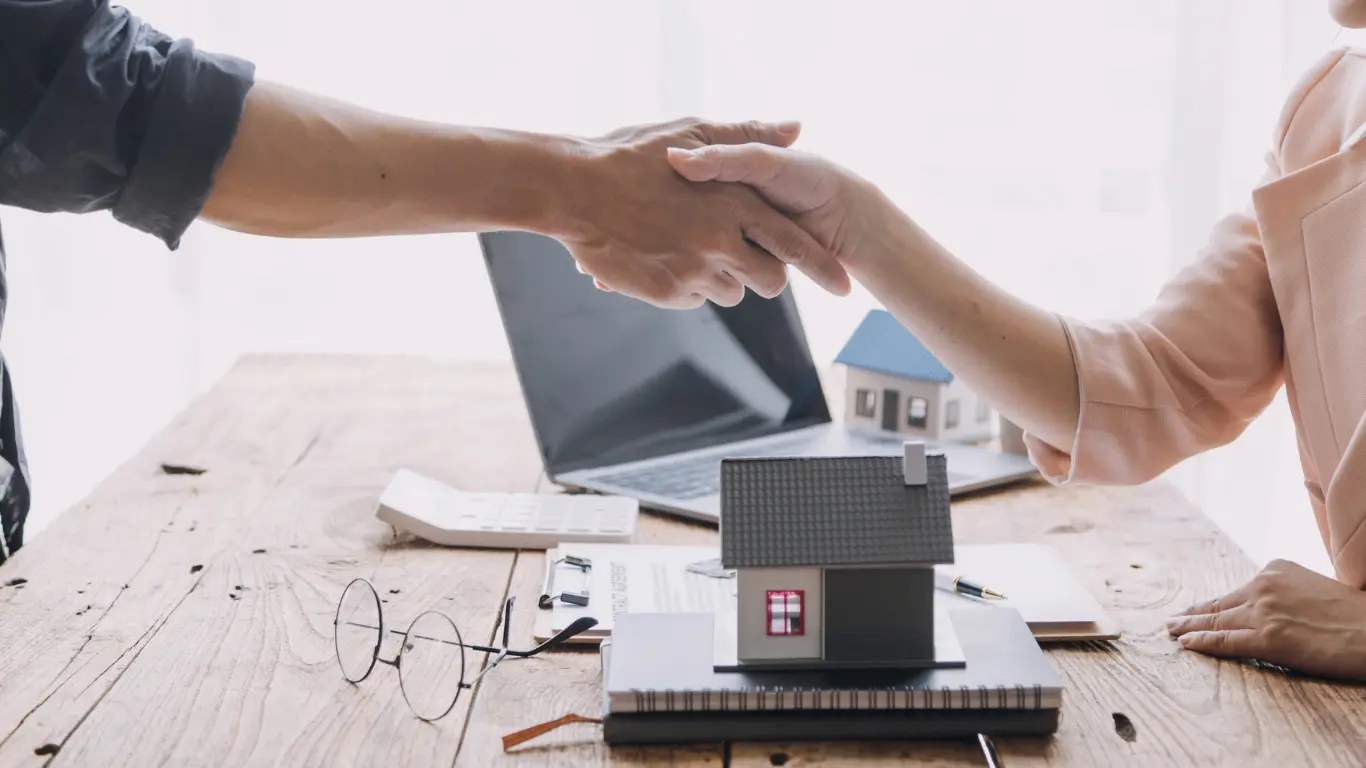 Duas pessoas apertando as mãos sobre uma mesa com uma maquete de casa, documentos e um notebook ao fundo, representando acordo ou negociação no mercado imobiliário.