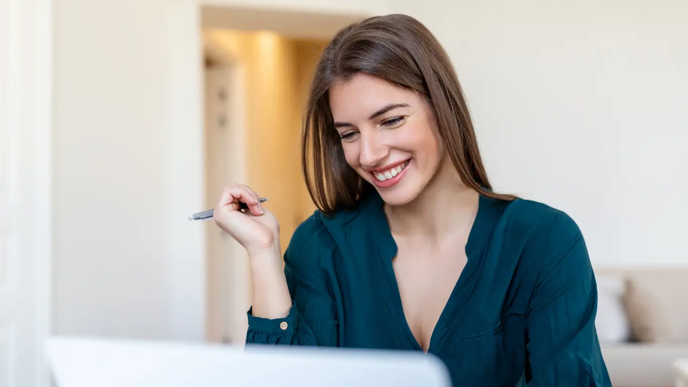 Mulher sorridente usando blusa verde, sentada em ambiente interno e olhando para um laptop, segurando uma caneta na mão.