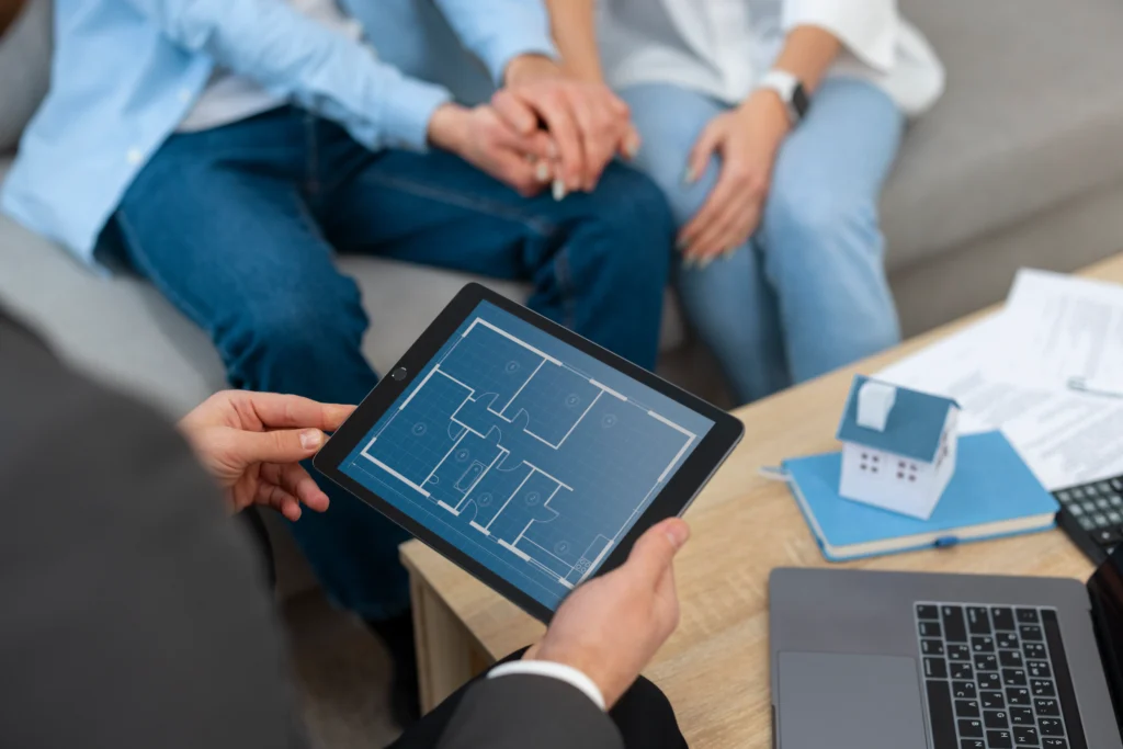 Otimização de vendas imobiliárias Otimização de vendas imobiliárias: a imagem mostra uma consulta imobiliária onde um corretor apresenta uma planta baixa em um tablet a um casal sentado em um sofá, enquanto uma miniatura de casa e documentos estão sobre uma mesa.