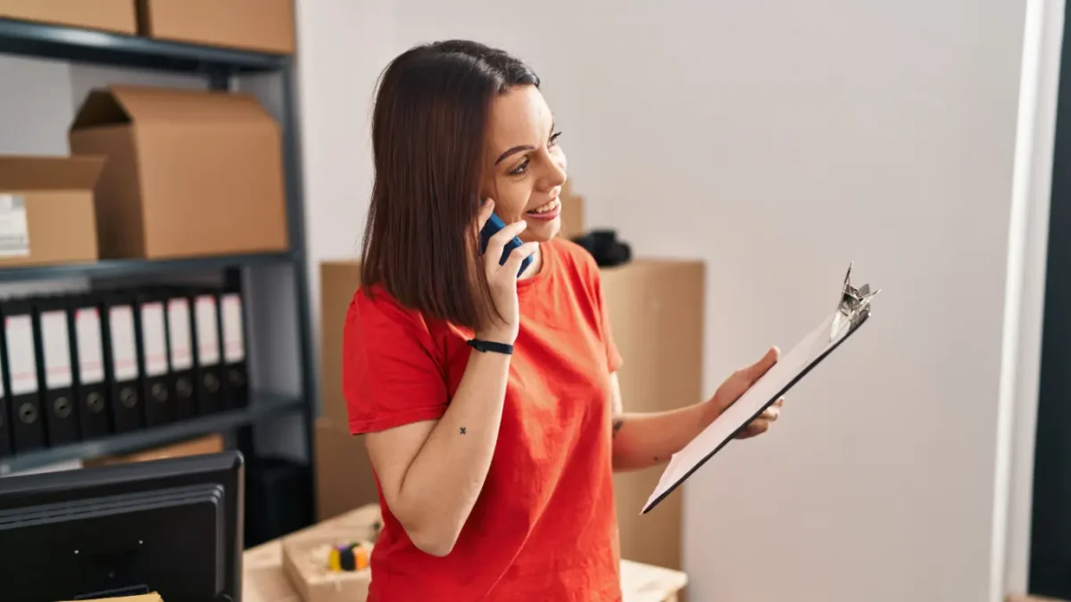 Mulher sorrindo enquanto fala ao telefone e consulta um documento em uma prancheta. Ela está em um ambiente de escritório com caixas e pastas ao fundo, sugerindo atividades relacionadas à gestão, organização ou atendimento no setor imobiliário.