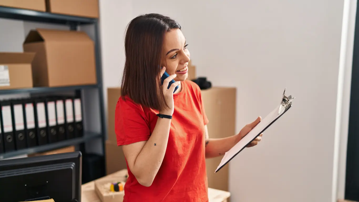 Mulher sorrindo enquanto fala ao telefone e consulta um documento em uma prancheta. Ela está em um ambiente de escritório com caixas e pastas ao fundo, sugerindo atividades relacionadas à gestão, organização ou atendimento no setor imobiliário.