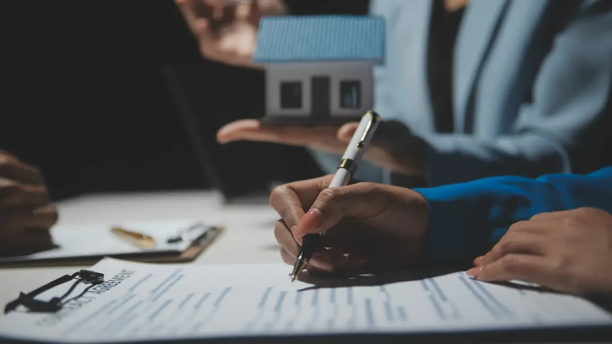 Pessoa assinando documentos imobiliários durante negociação, enquanto corretor apresenta uma maquete de casa, simbolizando compra, venda ou regularização de imóvel.