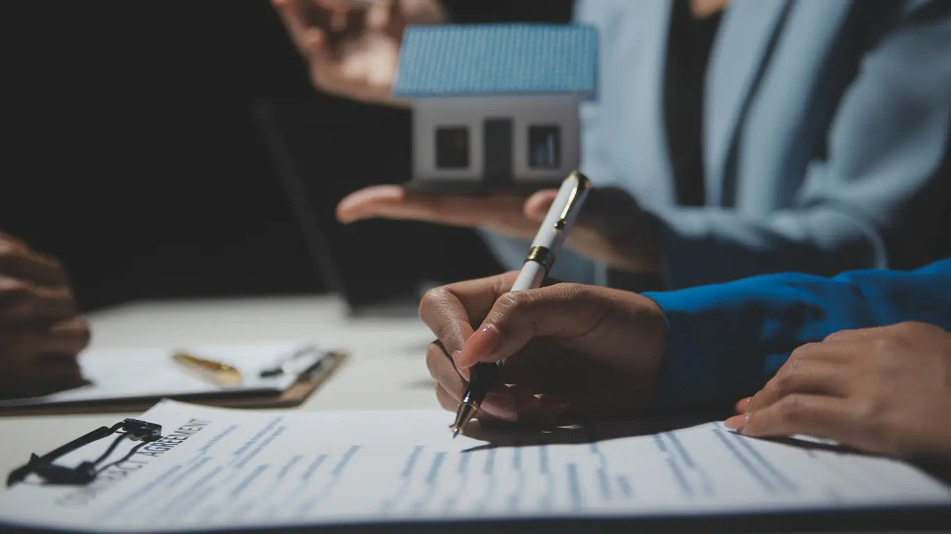 Pessoa assinando documentos imobiliários durante negociação, enquanto corretor apresenta uma maquete de casa, simbolizando compra, venda ou regularização de imóvel.