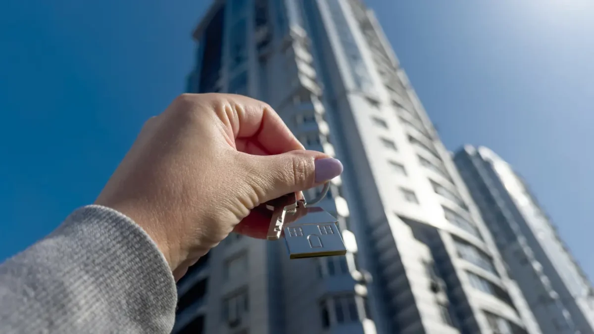 Mão segurando um chaveiro em formato de casa com chaves em primeiro plano, tendo ao fundo um edifício residencial alto sob céu azul. A imagem remete à compra, venda ou conquista de um imóvel e ao mercado imobiliário.