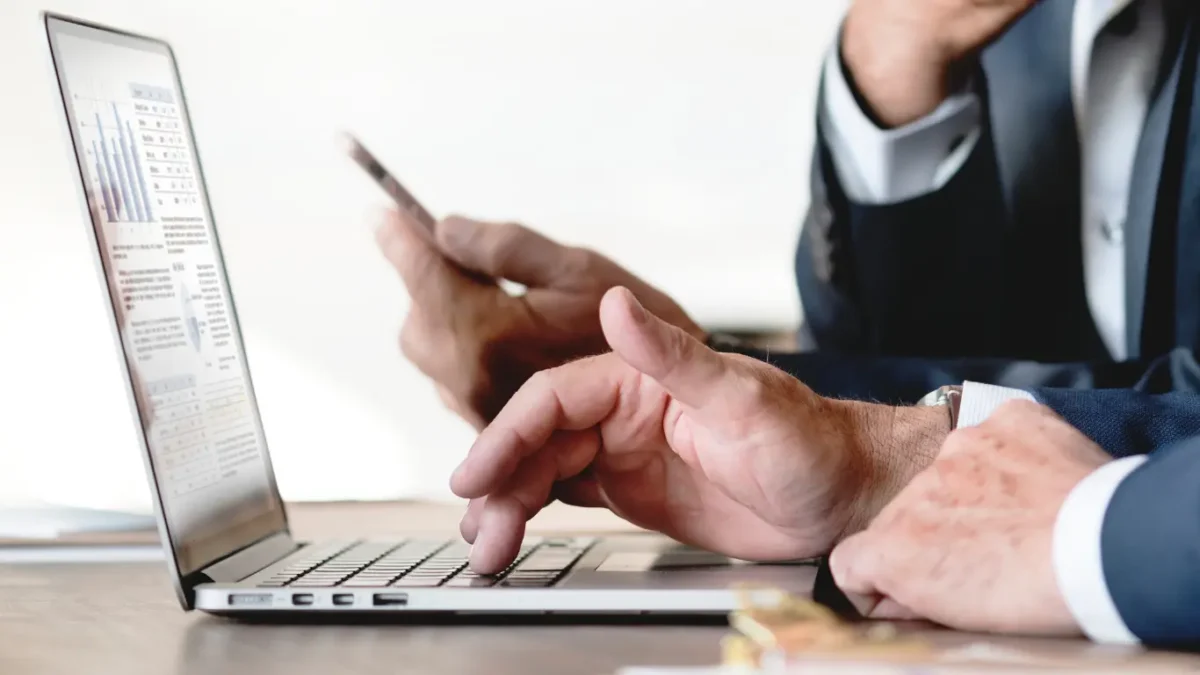 Plataformas para Corretores: A imagem em close-up mostra dois homens em terno trabalhando juntos em um ambiente profissional, focados em um laptop com gráficos e dados.