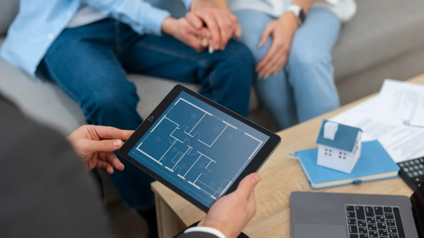 Consultor imobiliário apresenta a planta baixa de um imóvel em um tablet para um casal durante um atendimento. Sobre a mesa há uma maquete de casa, documentos e um notebook, representando a digitalização e a inovação no mercado imobiliário.