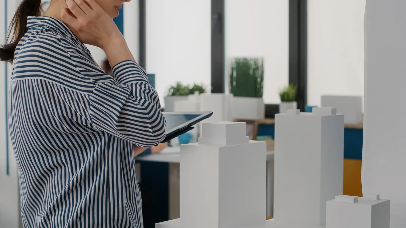 Mulher segurando um tablet e analisando maquetes de edifícios em um ambiente corporativo. A cena remete ao planejamento e à gestão de empreendimentos imobiliários, com modelos de prédios em destaque sobre uma mesa.