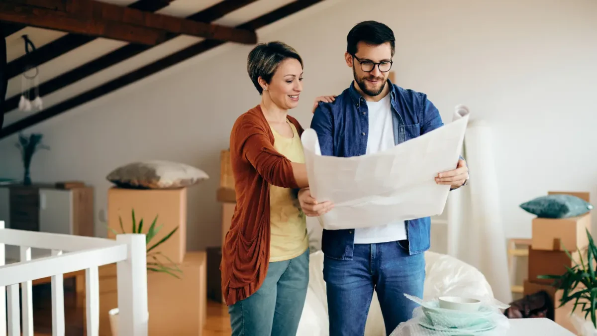 Casal em um imóvel durante uma mudança, analisando uma planta baixa ou projeto da casa. Ao redor, há caixas de papelão, móveis e objetos embalados, sugerindo planejamento da nova residência ou processo de compra e mudança para um novo lar.