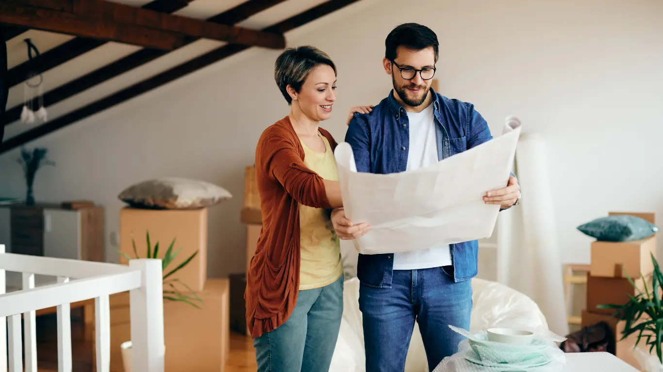 Casal em um imóvel durante uma mudança, analisando uma planta baixa ou projeto da casa. Ao redor, há caixas de papelão, móveis e objetos embalados, sugerindo planejamento da nova residência ou processo de compra e mudança para um novo lar.