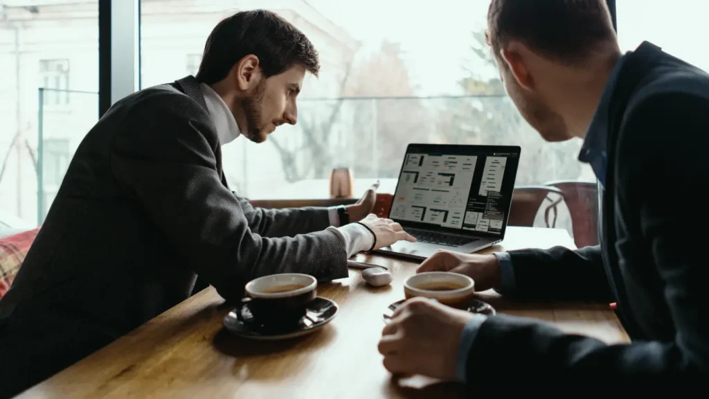 Plataformas para Corretores: Imagem mostra dois homens sentados em uma mesa, ambos com uma xícara de café a frente. Há uma computador onde um deles está digitando no teclado e o outro está olhando. Há uma janela de vidro ao fundo da imagem.