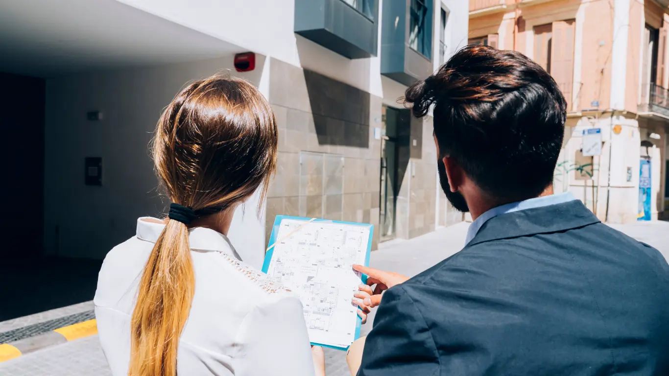 Corretor imobiliário apresenta a planta de um empreendimento a uma cliente em frente ao edifício, ilustrando o processo de geoanálise e avaliação de imóveis para compra ou investimento.