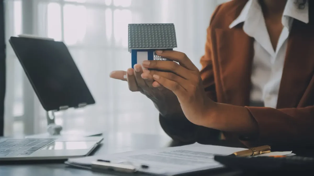 Pessoa em um ambiente de escritório segura uma maquete de casa com as duas mãos, ao lado de um notebook e documentos sobre a mesa. A imagem representa planejamento, análise e investimento em terrenos e empreendimentos imobiliários.