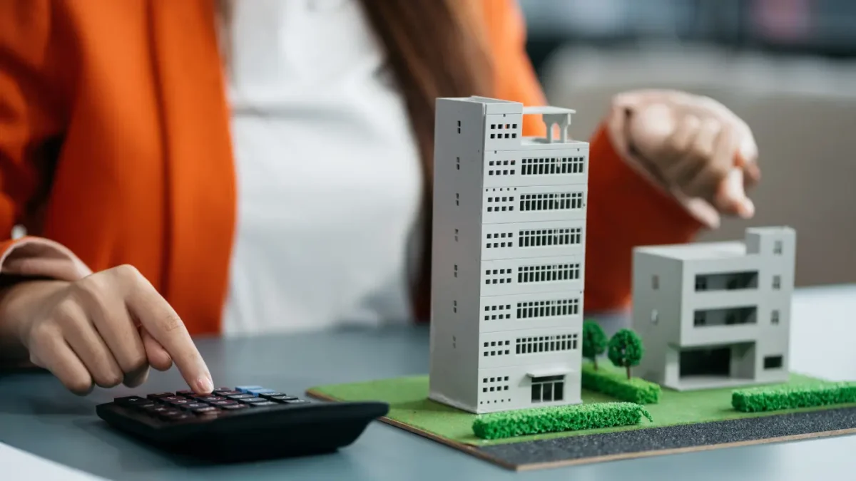 Pessoa utilizando uma calculadora ao lado de uma maquete de edifícios residenciais, representando análise de custos, investimentos e planejamento no mercado imobiliário.
