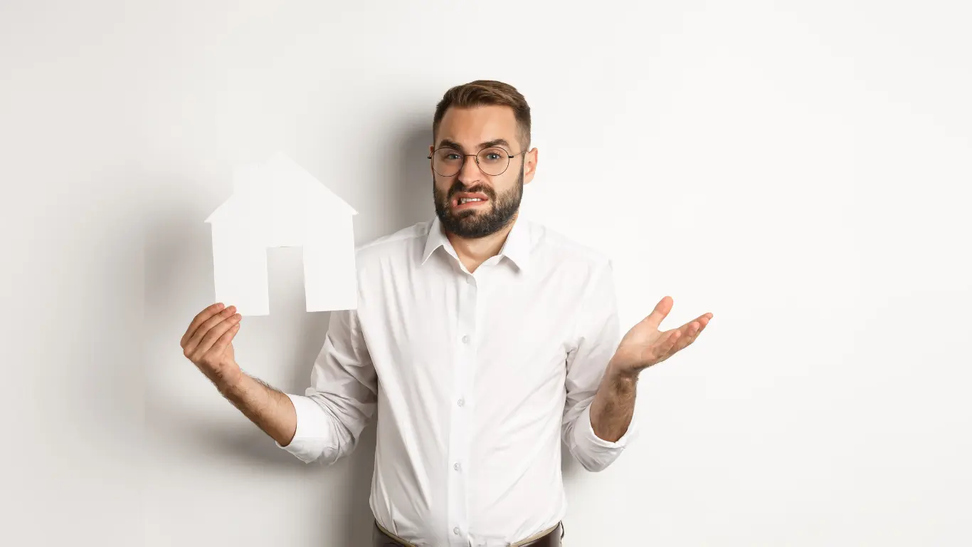 Homem de camisa branca e óculos segura um recorte de papel em formato de casa enquanto faz uma expressão de dúvida e levanta a outra mão. A imagem, com fundo branco e composição minimalista, representa incertezas relacionadas ao mercado imobiliário, compra ou gestão de imóveis.