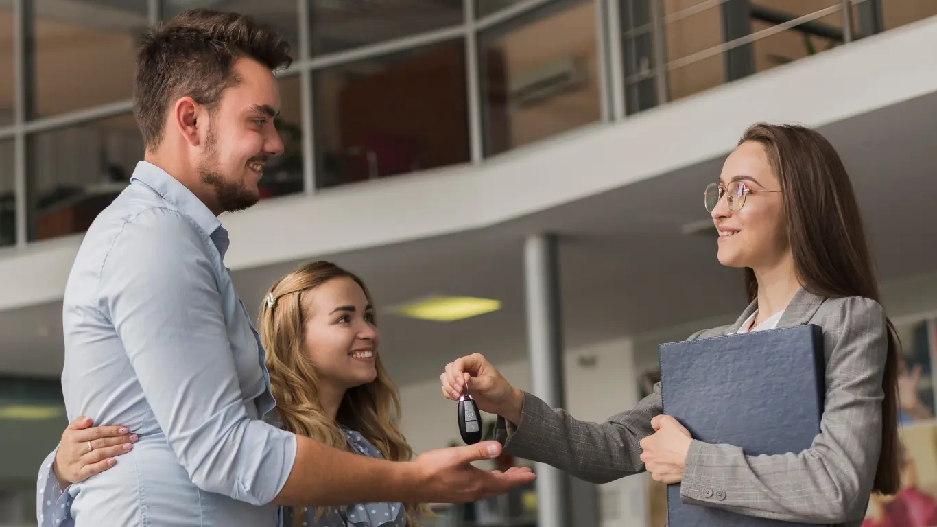 Corretora entrega as chaves de um novo imóvel a um jovem casal, que aparece sorrindo ao lado dela em um ambiente corporativo. A cena representa a conclusão da jornada de compra do imóvel e a realização do sonho da casa própria.