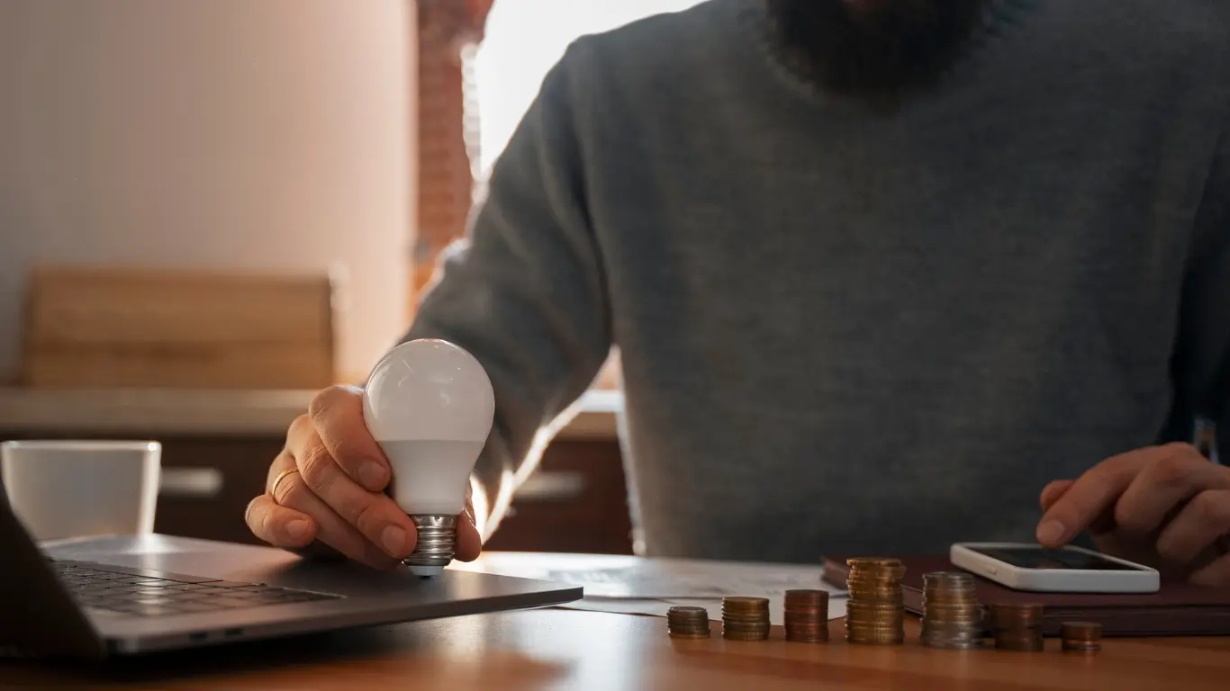 Pessoa segurando uma lâmpada LED sobre uma mesa, ao lado de um notebook, celular e moedas, representando planejamento financeiro, economia e investimento.