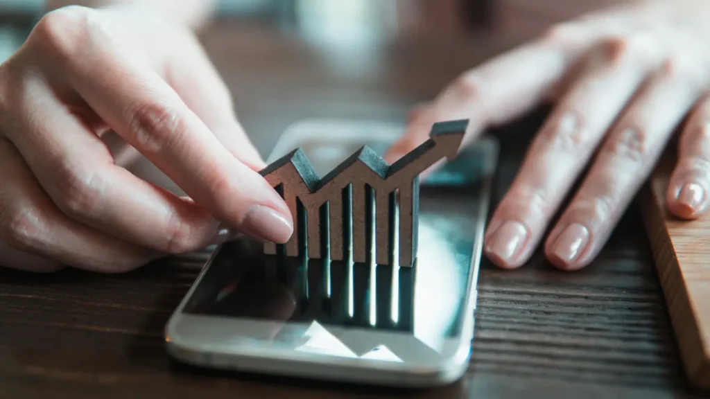 Canal de vendas imobiliário: Uma imagem de close-up mostra mãos femininas manipulando um gráfico de barras com seta ascendente sobre um smartphone, simbolizando crescimento e finanças digitais.