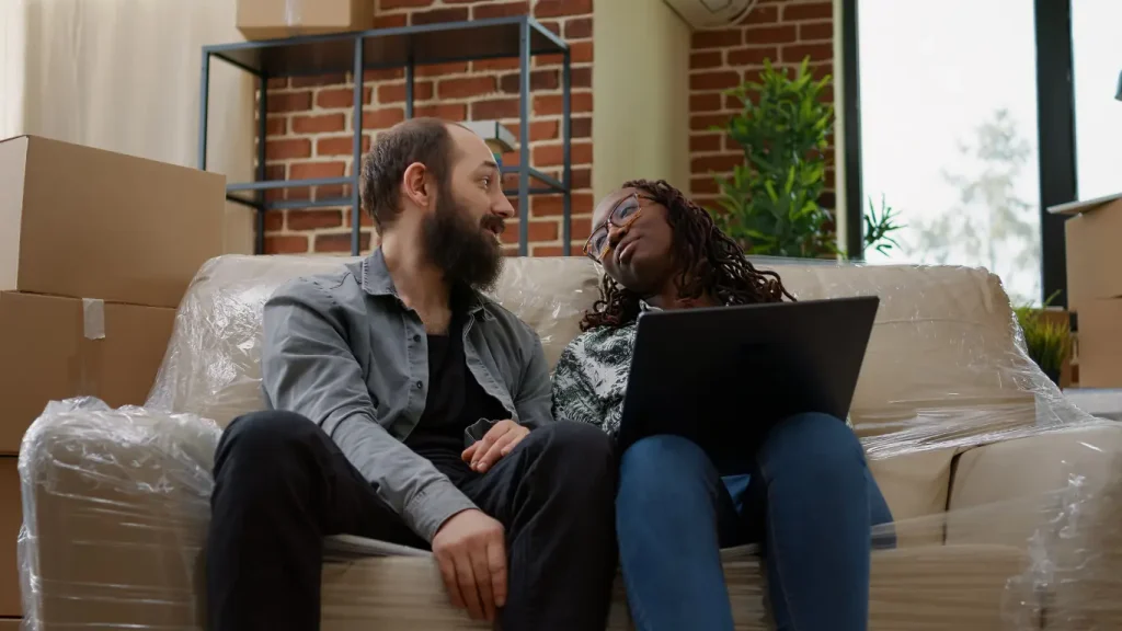 Go-to-market imobiliário: Um casal sentado no sofá, que está com uma capa de plástico. A mulher segura um notebook e o homem olhando para ela. 
